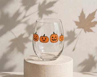 May include: A clear stemless wine glass featuring a horizontal row of four orange jack-o'-lantern pumpkins with black facial features, including eyes, noses, and smiling mouths, printed around the center of the glass.