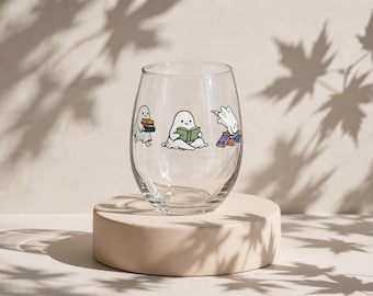 May include: A clear stemless wine glass featuring three small white ghost illustrations. One ghost carries a stack of books, another sits while reading an open book, and the third ghost leans over a small pile of colorful books.