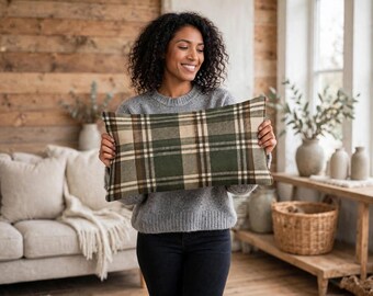 May include: A rectangular throw pillow featuring a plaid pattern in shades of forest green, cream, and dark brown. The pillow has a soft, woven texture and is held horizontally, displaying a classic grid design across its entire surface.