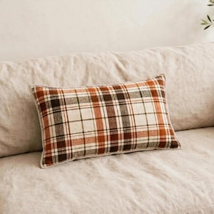 May include: A rectangular throw pillow featuring a plaid pattern with intersecting lines in shades of burnt orange, dark brown, olive green, and cream. The fabric has a visible woven texture, and the pillow is finished with a simple piped edge.