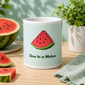 May include: A white ceramic mug with a light blue background featuring a graphic illustration of a red watermelon slice with black seeds and the dark green text One In a Melon printed underneath the watermelon graphic on the front side.