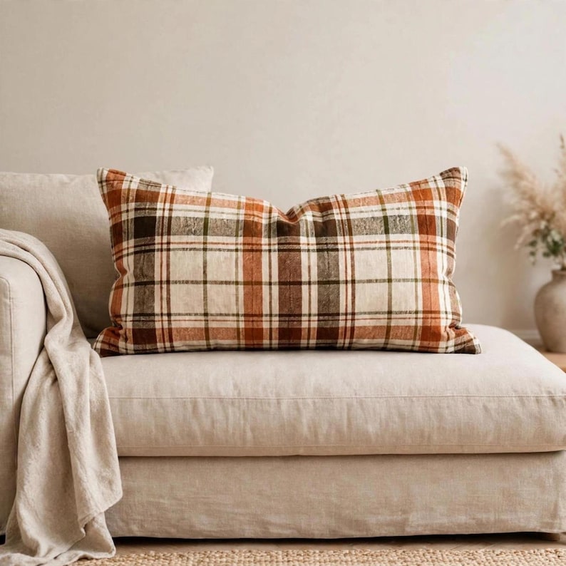May include: A rectangular throw pillow featuring a plaid pattern in shades of cream, burnt orange, brown, and dark green. The pillow has a woven texture and is positioned horizontally, displaying a classic grid design across its entire surface.