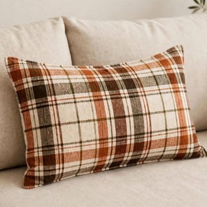 May include: A rectangular throw pillow featuring a plaid pattern with intersecting lines in shades of burnt orange, dark brown, and cream. The fabric has a visible woven texture, and the design consists of a repeating grid of horizontal and vertical stripes.