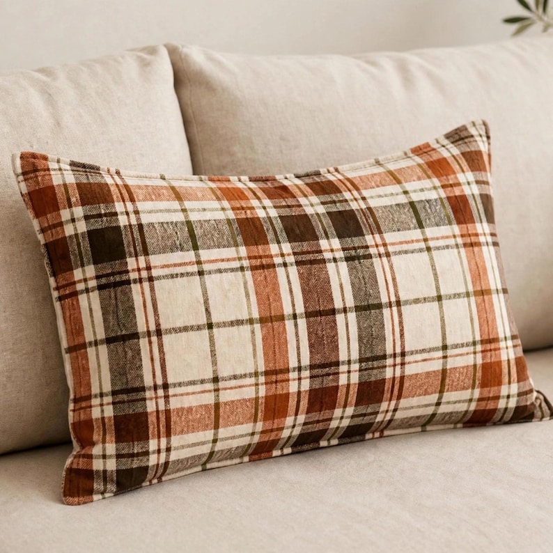 May include: A rectangular throw pillow featuring a plaid pattern with intersecting lines in shades of burnt orange, dark brown, and cream. The fabric has a visible woven texture, and the design consists of a repeating grid of horizontal and vertical stripes.