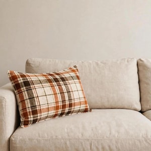 May include: A rectangular throw pillow featuring a plaid pattern in shades of cream, burnt orange, and dark brown. The pillow has a woven texture and is positioned on a light beige sofa. The design consists of intersecting horizontal and vertical lines.