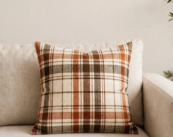 May include: A square throw pillow featuring a plaid pattern with intersecting lines in shades of cream, burnt orange, dark brown, and muted olive green. The fabric has a visible woven texture, and the pillow is plumped into a square shape.