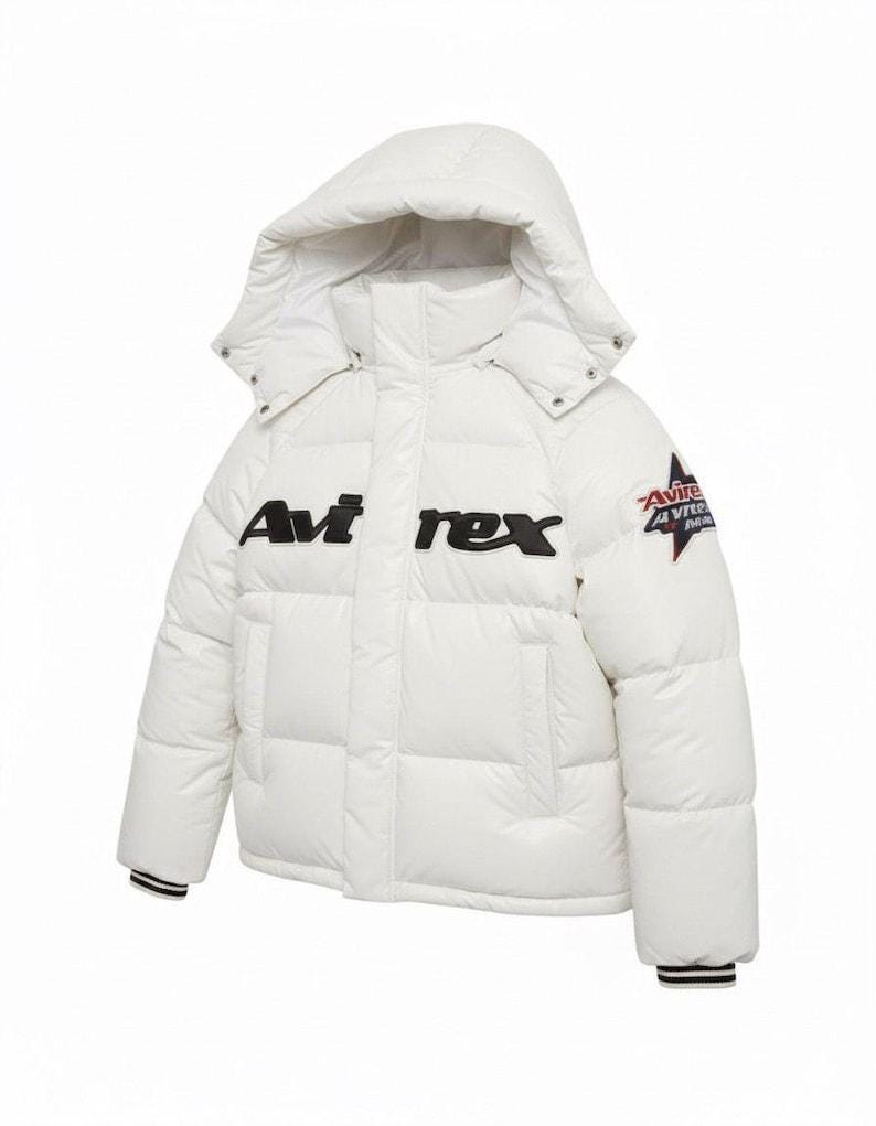 May include: A white puffer jacket with a hood. The jacket features the brand name "Avi rex" in black block letters across the chest and a star logo on the sleeve. The cuffs have black and white stripes.