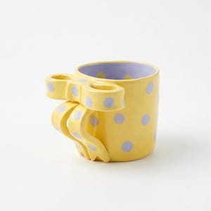 May include: A whimsical yellow ceramic mug with a bow-shaped handle and lavender interior. The mug is adorned with lavender polka dots. The handle is a unique design element, adding a playful touch to the drinkware.