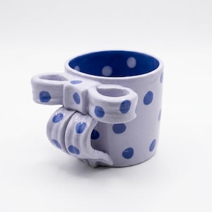 Könnte beinhalten: Eine lavendelfarbene Keramiktasse mit blauen Tupfen und blauem Innenraum. Der Griff ist wie eine Schleife geformt. Die Tasse ist ein dekorativer Gegenstand. Das Design der Tasse ist skurril und einzigartig.
