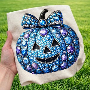 May include: A beige fabric tote bag featuring a large pumpkin graphic decorated with numerous blue and purple faceted rhinestones. The pumpkin has a black stem, a bow on top, and a carved jack-o'-lantern face with triangular eyes and a wide grin.