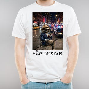 Peut inclure: Un t-shirt blanc avec une illustration d'un raton laveur portant un costume bleu, un fedora beige et des lunettes de soleil, assis sur une chaise dans une rue urbaine la nuit avec une boisson. Le texte "i live here now" est écrit en noir.