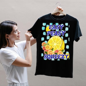 Puede incluir: Una camiseta negra de manga corta con un estampado gráfico de varias rodajas de cítricos, incluyendo limones y pomelos, sobre un fondo de cuadros azul claro. El texto "EASY PEASY SQUEEZY" está impreso en letras azules en negrita.