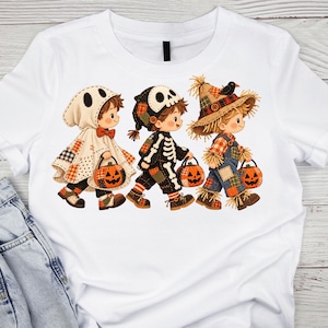 Op de afbeelding: Een wit T-shirt met korte mouwen en een afbeelding van drie kinderen in Halloween-kostuums. Eén kind draagt een spookcape, één een skeletpak en één een vogelverschrikker-outfit. Elk kind draagt een klein oranje pompoen-snoepemmertje.