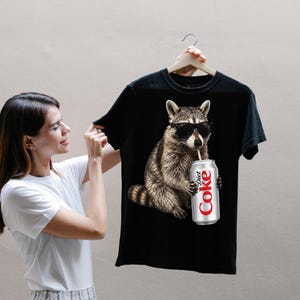 Puede incluir: Una camiseta negra de manga corta con un estampado gráfico de un mapache que lleva gafas de sol negras y sostiene una lata plateada de Diet Coke con una pajita. El mapache está representado con un estilo realista y pelaje detallado.