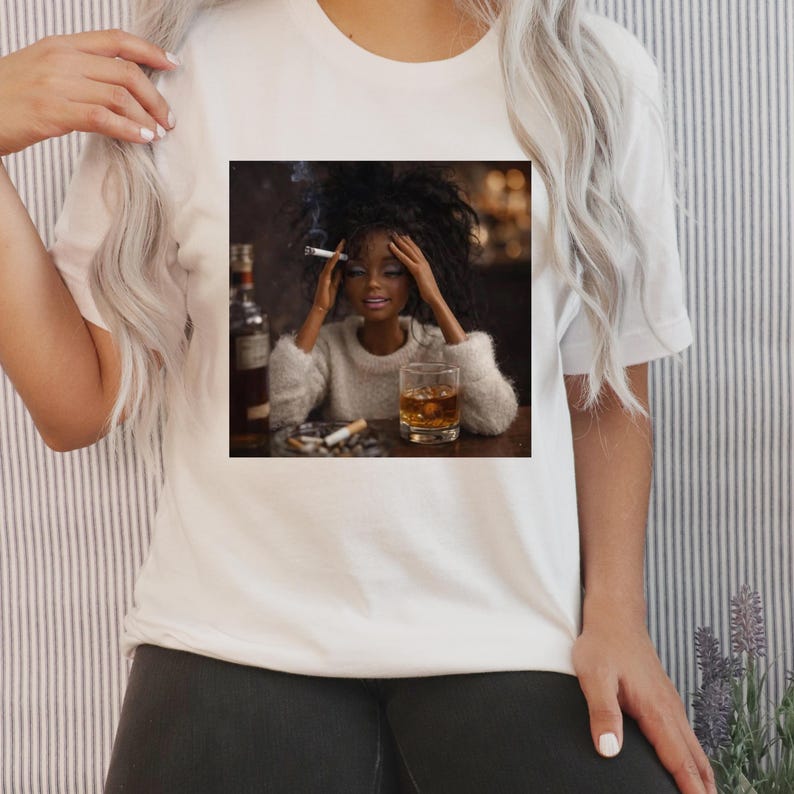 May include: White t-shirt featuring a square image of a doll with dark hair, holding a lit cigarette, and sitting at a table with a glass of whiskey and a bottle. The doll is wearing a fuzzy sweater. Cigarette butts are in the ashtray.