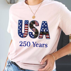 Pode incluir: Uma t-shirt de manga curta rosa claro com o texto USA em letras grandes com missangas e um padrão de bandeira vermelha, branca e azul, e o texto 250 Years bordado a azul por baixo. A t-shirt é usada com calças de ganga azuis e um colar dourado.