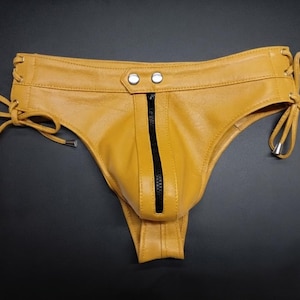 Może przedstawiać: Musztardowy skórzany jockstrap z pionowym czarnym zamkiem błyskawicznym z przodu, dwoma srebrnymi metalowymi zatrzaskami w pasie oraz regulowanymi sznurowaniami po bokach z metalowymi srebrnymi końcówkami.