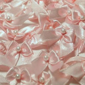 May include: Close-up of many small, light pink satin bows with a pearl bead in the center of each. The bows are arranged closely together, creating a textured, decorative pattern. Ideal for crafts and embellishments.