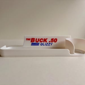 May include: A white plastic tray with a raised back and a small cup holder. The text "THE BUCK .50 GLIZZY" is printed on the back in red and blue. The tray is designed to hold items.
