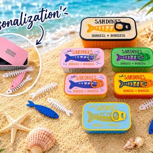 Op de afbeelding: Een verzameling rechthoekige blikjes met het label not SARDINES SKINLESS + BONELESS in diverse kleuren zoals oranje, roze, groen, geel en blauw. Elk blikje heeft een visafbeelding en een deksel met lipje. Eén roze blikje is gepersonaliseerd met JENNI.