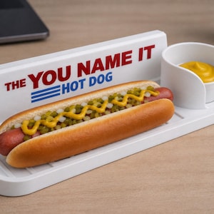 May include: A white plastic hot dog serving tray with a built-in condiment cup and a back panel featuring the red and blue text THE YOU NAME IT HOT DOG. A hot dog in a bun topped with yellow mustard and green relish sits on the tray base.