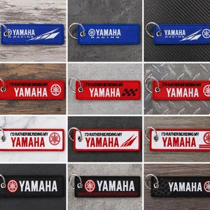Op de afbeelding: Collectie Yamaha sleutelhangers in verschillende kleuren, waaronder blauw, rood, wit en zwart. Elke sleutelhanger heeft het Yamaha logo en tekst, sommige met race-thema ontwerpen. De sleutelhangers zijn rechthoekig met een metalen ring.