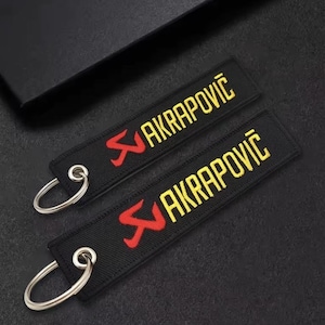 Peut inclure: Deux porte-clés noirs avec le mot "AKRAPOVIČ" en jaune et un logo rouge. Chaque porte-clés est muni d'un anneau métallique argenté. Les porte-clés sont sur une surface sombre.