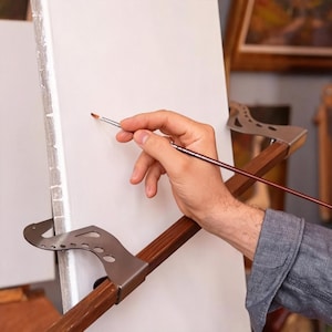 Könnte beinhalten: Eine weiße Leinwand wird von zwei silbernen Metallklammern gehalten, die an einer horizontalen Holzstange befestigt sind. Ein dünner Pinsel ist gegen die Oberfläche der Leinwand gerichtet. Die Metallklammern haben ein geschwungenes Design mit Aussparungen.