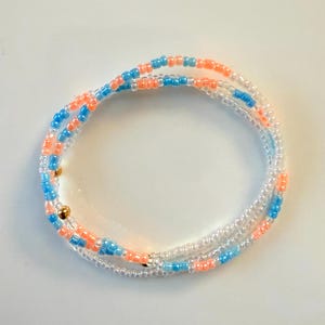 May include: A multi-strand beaded bracelet featuring small, round glass beads in alternating shades of light blue, peach, and translucent white. The design includes two small, polished gold-toned metallic beads interspersed among the colorful glass beads.