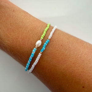 May include: Two beaded bracelets featuring small round beads in shades of lime green, bright blue, and white. One bracelet includes a single white oval freshwater pearl accent, while both bracelets are accented with tiny gold-toned metal beads.
