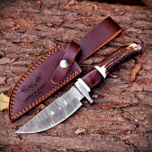 May include: A fixed-blade knife with a patterned Damascus steel blade and a textured brown antler handle with white spacers. The knife includes a dark brown leather sheath featuring a snap closure, orange stitching, and the text ANNA & JOHN 11.04.2025.