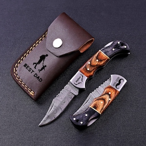 May include: A brown leather sheath with a snap closure, featuring a black silhouette of a parent and child and the text BEST DAD, alongside two folding pocket knives with Damascus steel blades and handles made of dark wood and black horn accents.