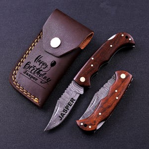 May include: A brown leather sheath with a snap closure and the text Happy Birthday Jasper, alongside two folding pocket knives with dark wood handles and Damascus steel blades. One blade is engraved with the name JASPER in capital letters.