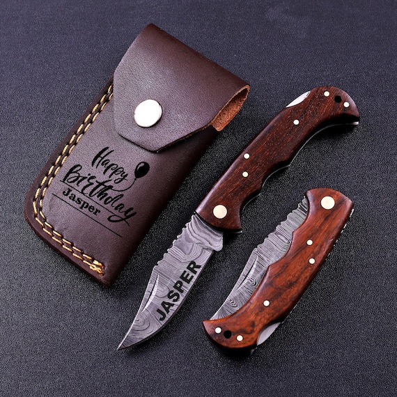 A hand-forged Damascus steel folding EDC tool paired with a custom leather sheath — the kind of rugged, tactile carry piece that looks like it belongs in a blacksmith's portfolio.