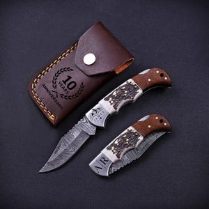 May include: Two folding pocket knives with Damascus steel blades and handles featuring wood and textured bone inlays. One knife is open, engraved with "JAMES" and a mountain icon. The other is closed, engraved with "A|R". A brown leather sheath says "10 YEARS ANNIVERSARY".