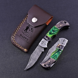 May include: A brown leather sheath with a snap closure and the text HAPPY ANNIVERSARY with a couple illustration, alongside two folding pocket knives featuring green and dark wood handles, metal bolsters, and patterned steel blades.