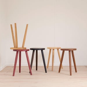 May include: A selection of wooden stools in different colours and finishes. One stool is inverted, showing its natural wood grain. Other stools are painted in red, black, light wood, and dark wood tones. The stools have three legs and round seats.