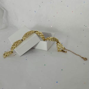 May include: A gold-tone bracelet featuring a series of interconnected, textured, leaf-shaped links. The bracelet includes a thin chain extension with a small circular clasp at the end, resting partially inside and over a small, open white square box.