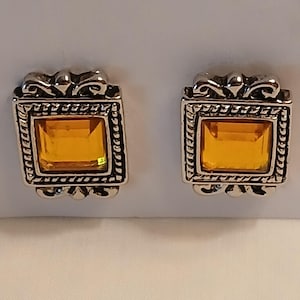 May include: A pair of silver-toned square stud earrings. Each earring features a rectangular amber-colored stone set within a decorative frame with a rope-like border and ornate top design.