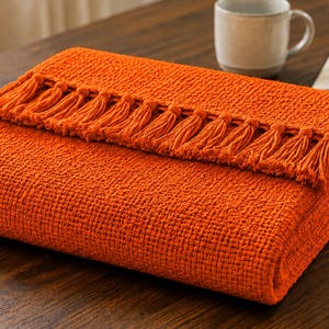 Könnte beinhalten: Eine leuchtend orangefarbene Wohndecke mit einer strukturierten Waffelstrickoptik und einer Reihe passender orangefarbener Quasten entlang der gefalteten Kante. Die Decke ist ordentlich rechteckig gefaltet und zeigt das gleichmäßige Webmuster.