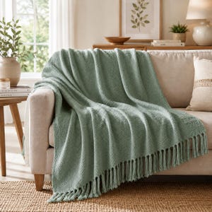 May include: A sage green throw blanket with a textured, woven pattern and long fringe detailing along the bottom edge. The blanket is draped over the arm and seat of a neutral-colored sofa, showcasing its soft, tactile surface and consistent color.