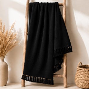 May include: A solid black throw blanket with a textured, fleece-like appearance. The blanket features long, matching black fringe tassels along the bottom edge and is draped over a wooden ladder frame to display its length and drape.