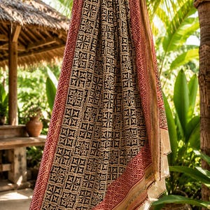 Puede incluir: Un textil estampado con un diseño negro, beige y rojo, drapeado sobre una viga de madera. La tela presenta un patrón floral y geométrico repetitivo. Los bordes están adornados con un borde rojo y detalles dorados. El fondo incluye un exuberante follaje verde.