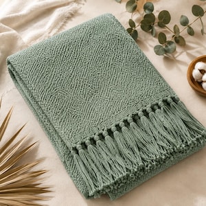 May include: A sage green throw blanket featuring a textured, raised geometric pattern across the surface. The blanket is finished with a row of long, matching sage green fringe tassels along one edge, creating a decorative border detail.