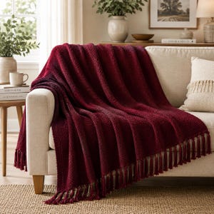 May include: A deep burgundy throw blanket with a textured, ribbed knit pattern and long, matching tassels along the bottom edge. The blanket is draped over a light-colored sofa, showcasing its thick, soft material and rich, dark red color.