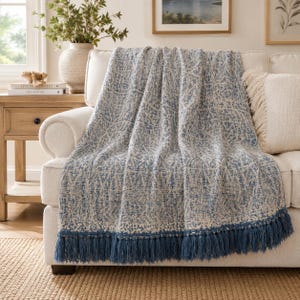 May include: A woven throw blanket featuring a blue and cream geometric pattern with a textured finish. The blanket is draped over a sofa and is finished with a long, blue fringe along the bottom edge, adding a decorative element to the textile.