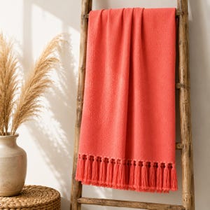 May include: A solid coral-colored throw blanket with a textured surface and a row of matching fringe tassels along the bottom edge. The rectangular blanket is draped over a wooden ladder, displaying its soft fabric and uniform vibrant color.