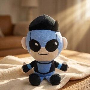 May include: A plush doll in a superhero costume. The doll has a light blue head and body with black accents, large black eyes, and a black cap. It is sitting on a cream-colored blanket.