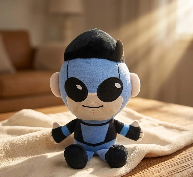 May include: A plush doll in a superhero costume. The doll has a light blue head and body with black accents, large black eyes, and a black cap. It is sitting on a cream-colored blanket.