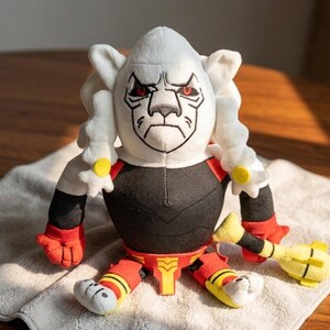 May include: A plush toy featuring a white head with a stern expression and red eyes. The toy has a black torso, red and yellow accents, and a yellow club. The toy is sitting on a white towel.